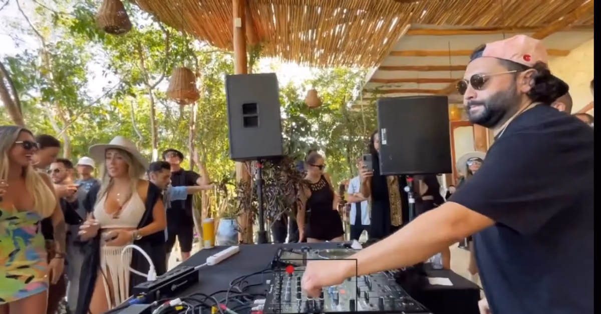 A DJ in sunglasses mixes music outdoors as a lively crowd dances and mingles under a bamboo canopy with trees behind.