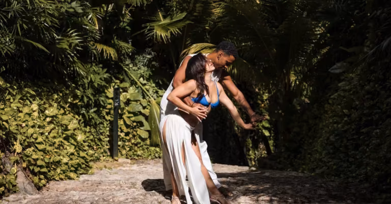 A couple dances on a stone path amid lush greenery. She wears a white skirt and blue bikini top; he wears white pants in dappled sun.