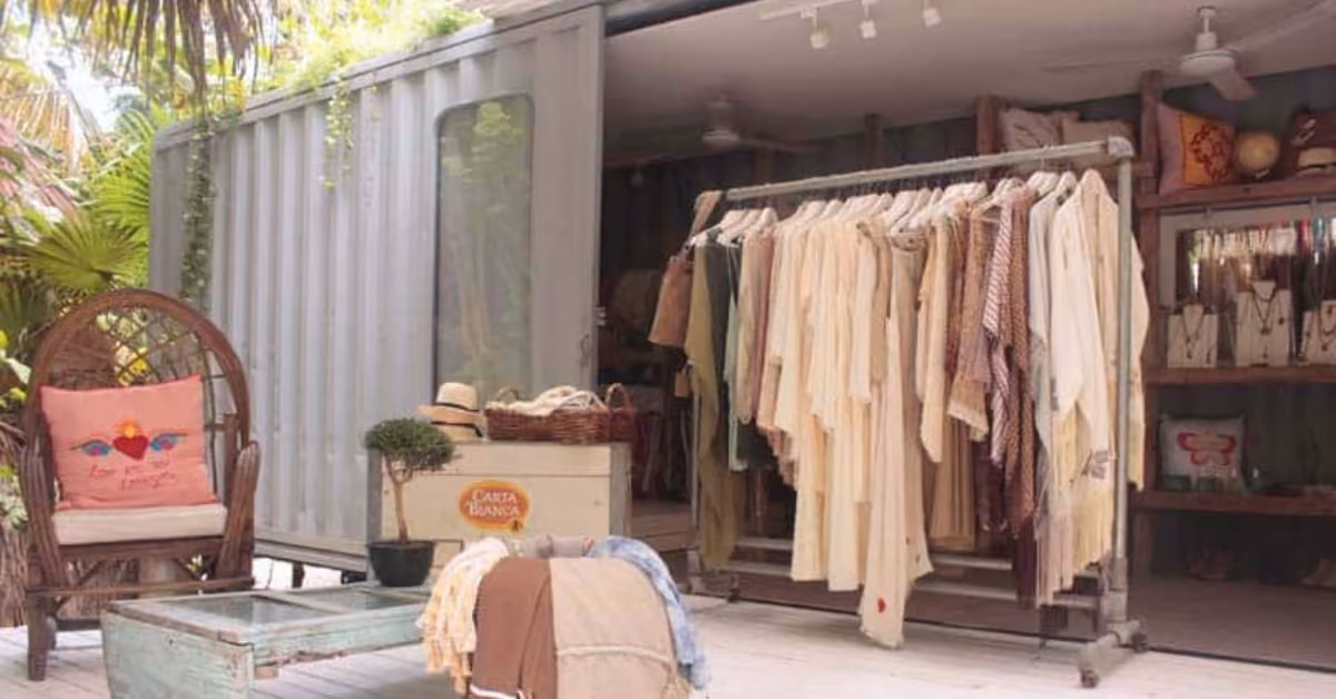Boutique in a shipping container with racks of neutral clothes, hats, jewelry, wicker chair, crates, and lush tropical plants.