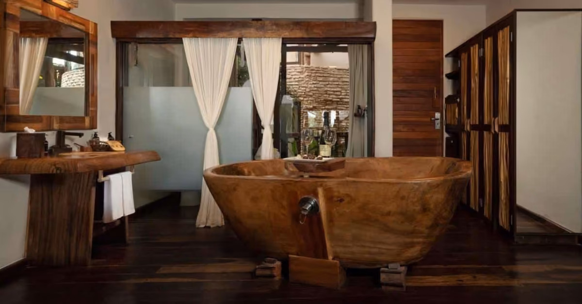 Rustic bathroom with large wooden tub, sink, and cupboards. White curtains cover the window; dark wood tones throughout.