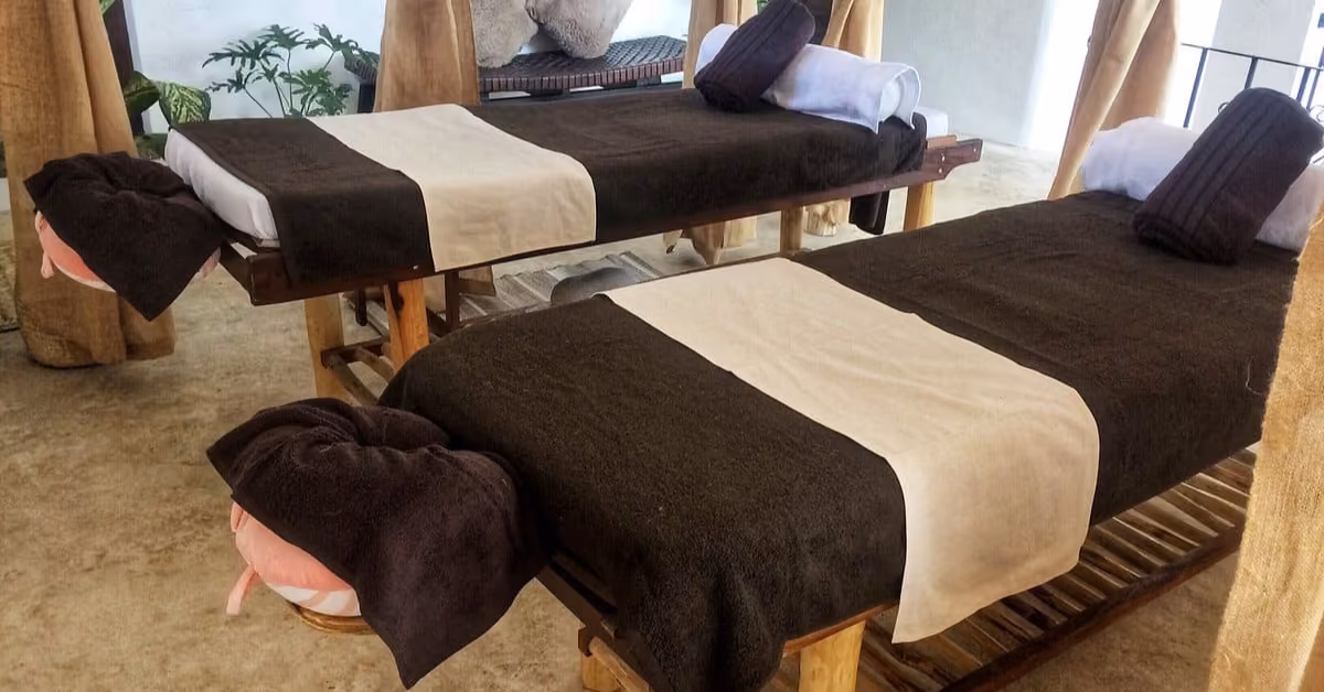 Two wooden massage tables with brown and beige linens are side by side in a serene spa room, each with towels and face cradles.