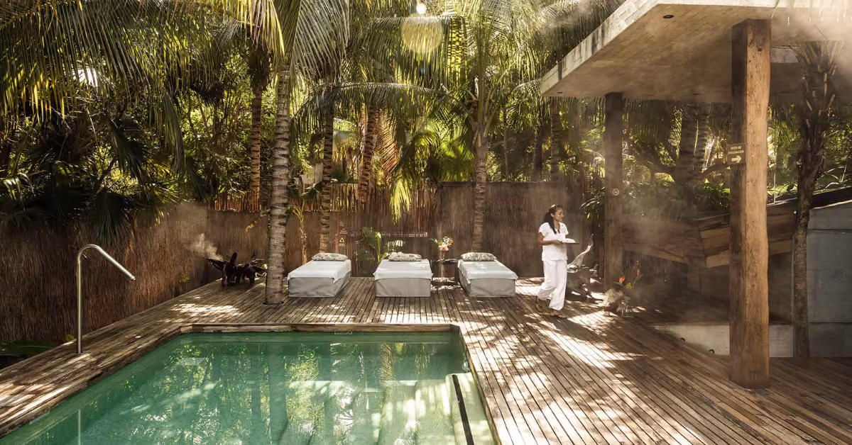 A peaceful outdoor spa with three massage beds by a pool, amid tropical trees. A person in white carries towels on the deck.