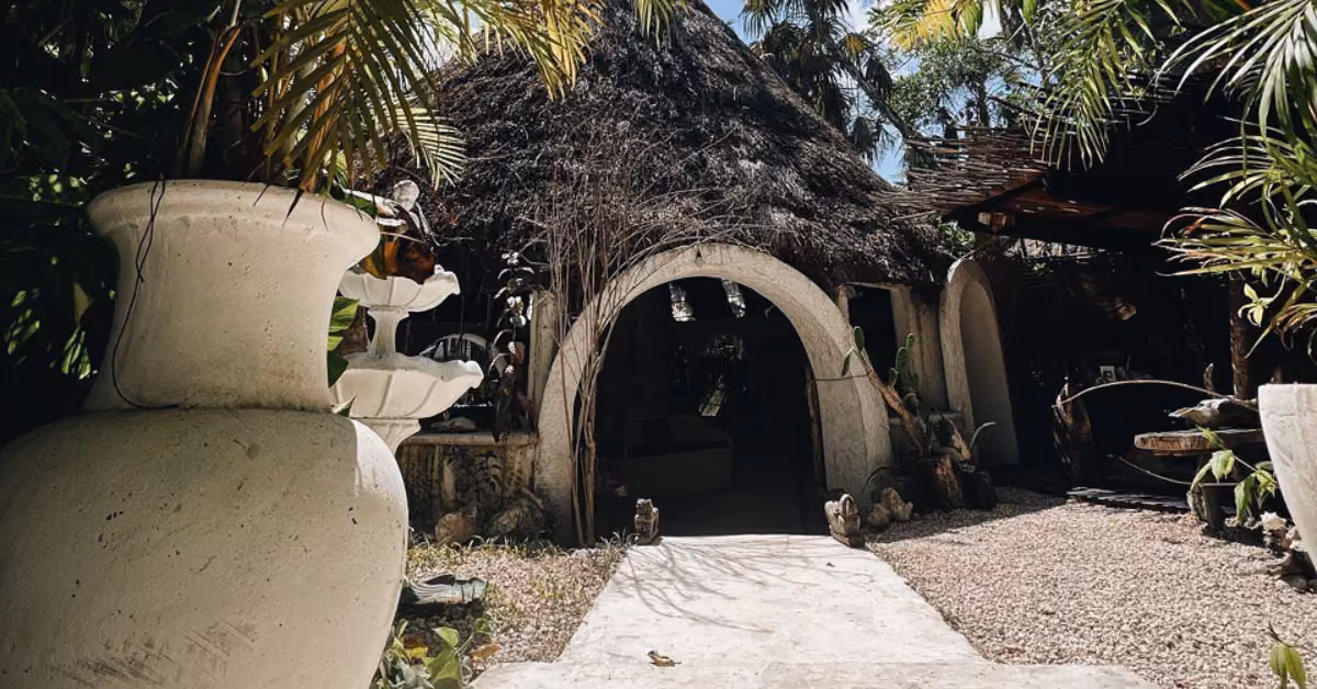 A rustic round hut with a thatched roof and arched entry sits amid lush greenery and white planters in bright sunlight.