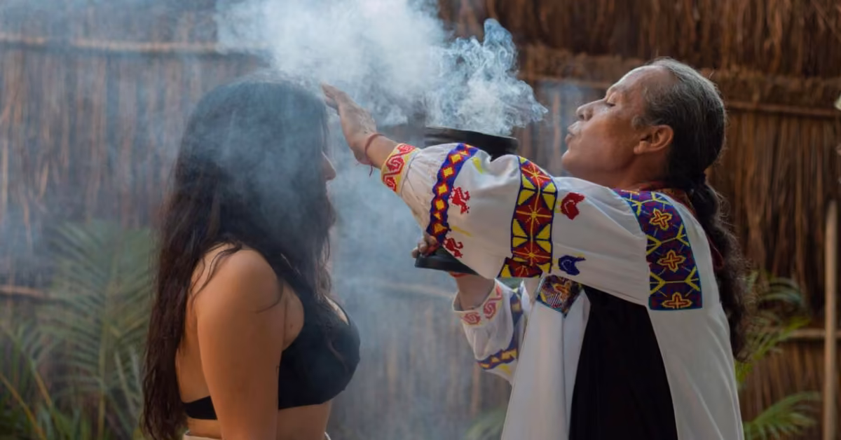 A person in traditional clothing performs a smoke ritual before someone in black, surrounded by plants and thatched walls.
