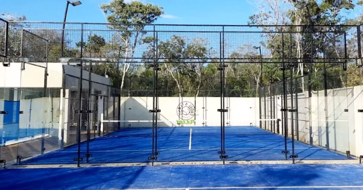 Blue-floored outdoor padel court with glass walls, black fencing, and white lines; trees and sky in the background.