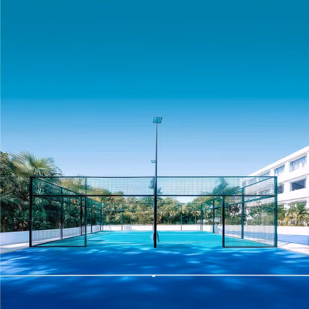 Empty blue padel tennis court with netting, palm trees, and modern buildings under a clear sky; tree shadows on the ground.