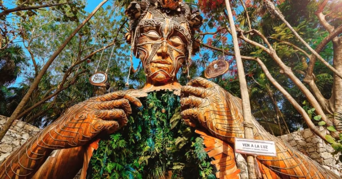 Wooden human sculpture with detailed carvings and greenery on its chest, surrounded by trees and a VEN A LA LUZ sign nearby.