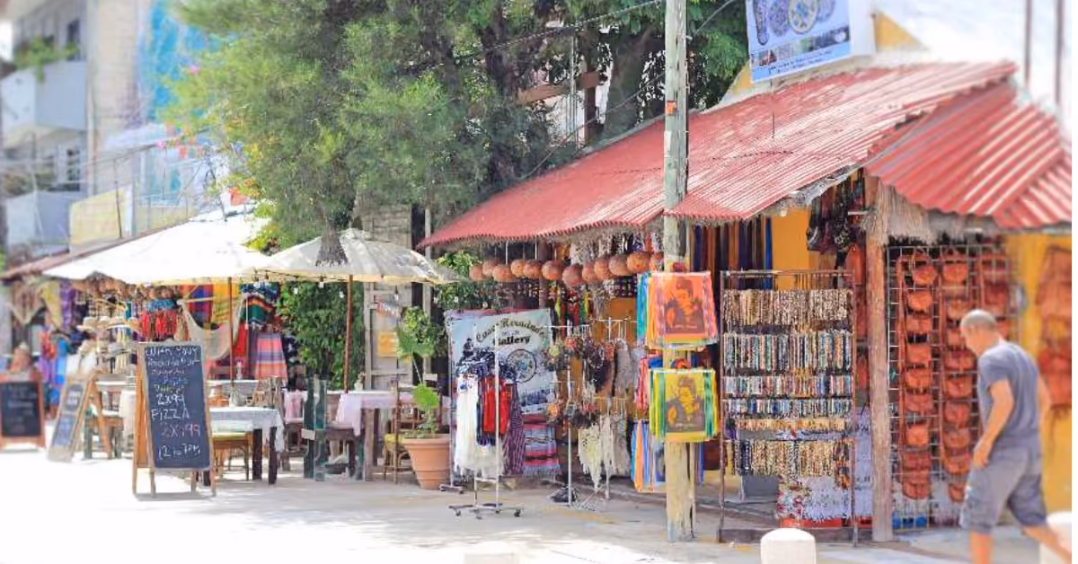 Downtown Tulum street market with colorful stalls, shaded by trees and umbrellas. A man passes a chalkboard sign for pizza.