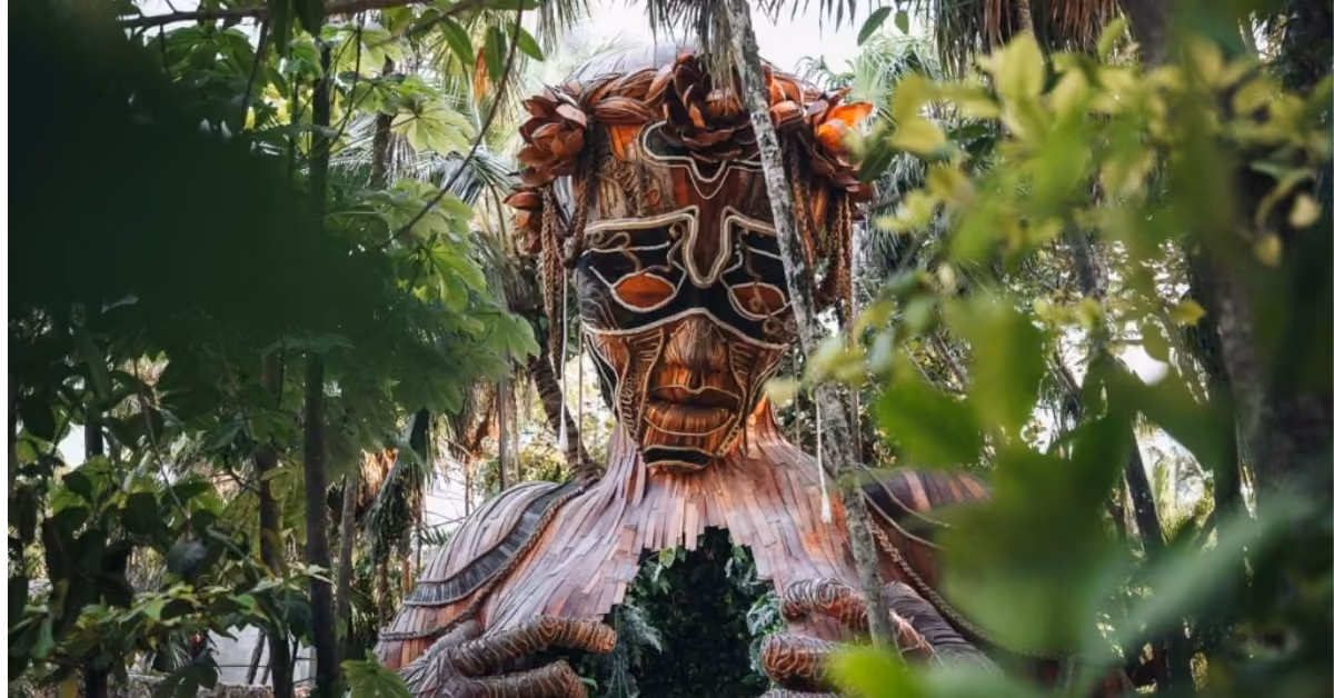 A carved wooden human sculpture stands among lush green foliage in Tulum, its chest opened to reveal plants inside.