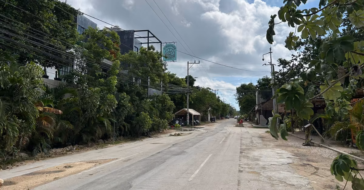 An empty street in La Veleta Tulum is lined with trees and buildings under a partly cloudy sky, with a sign above the road.