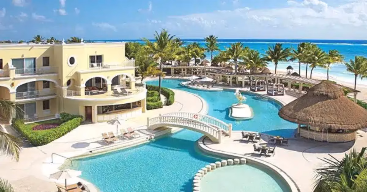 Tropical resort with a large pool, palm trees, thatched bar, lounge chairs, and yellow buildings beside a sandy beach and blue ocean.