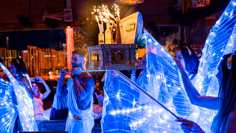 Performers in lit angel wings parade a bottle chest with sparklers through Ilios
