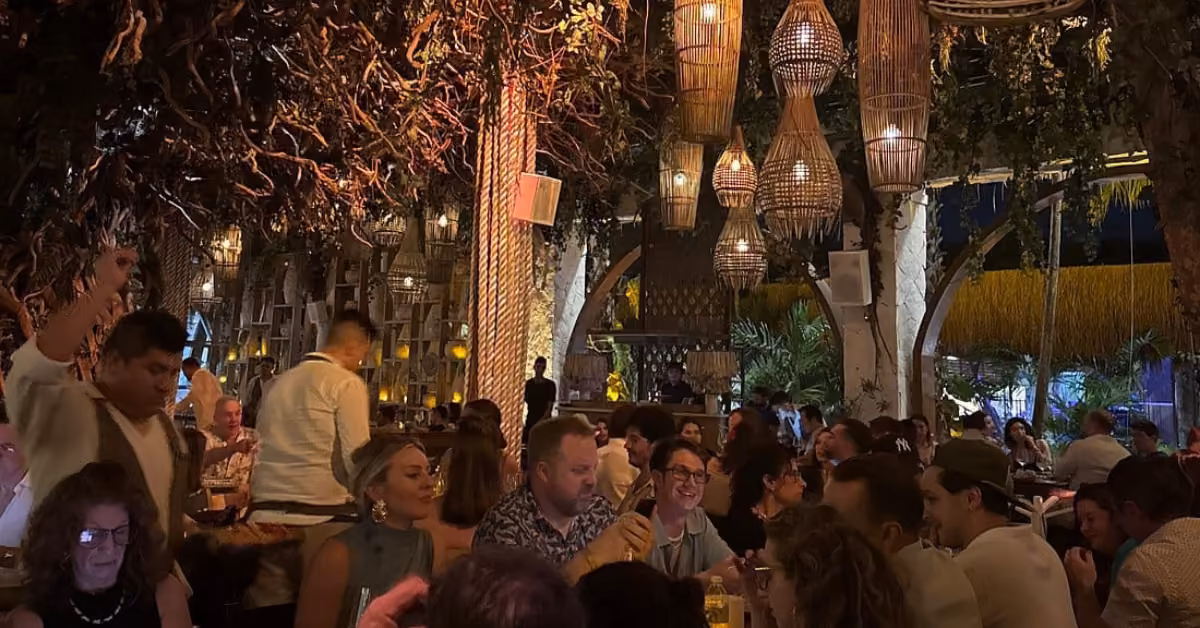 Ilios Restaurant glows with warm lighting, wicker lamps, and lush plants as groups dine in a vibrant, cozy setting.