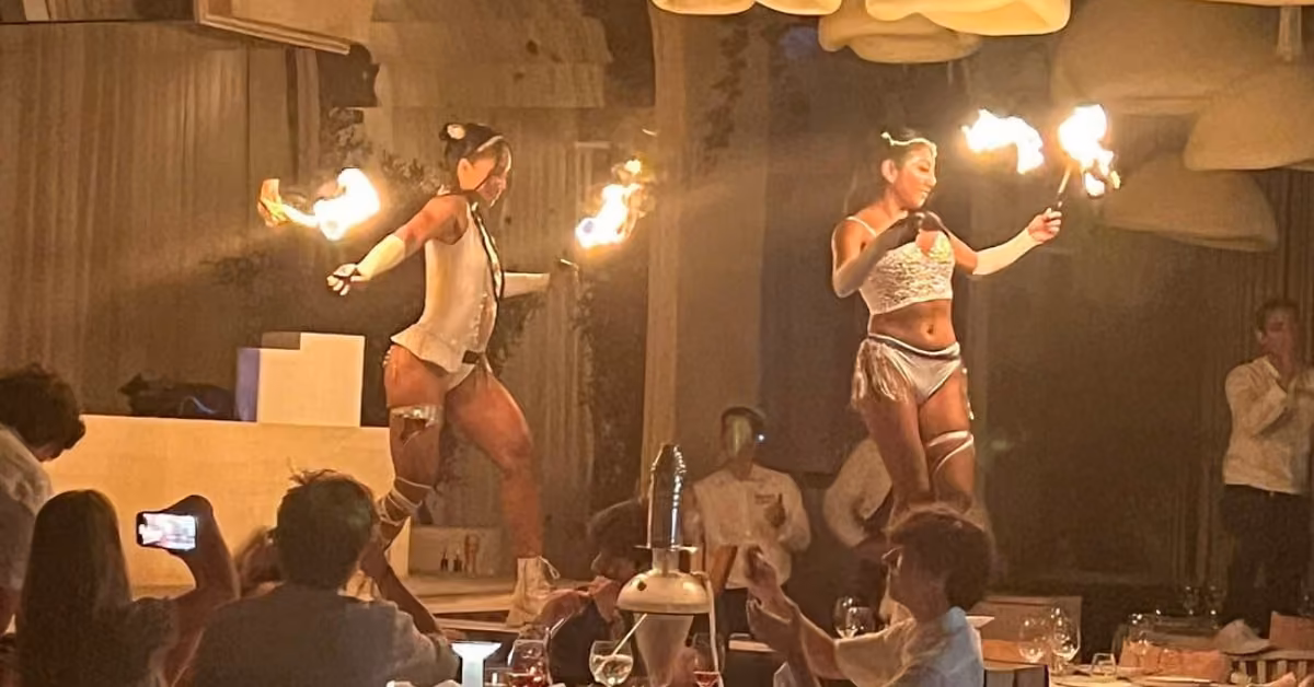 Two women in white costumes perform a torch dance on stage at a restaurant as diners watch and take photos.