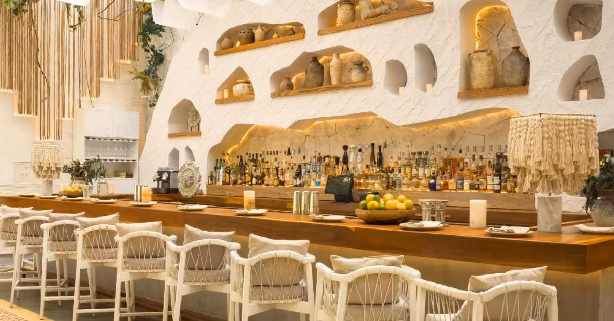 Ilios Restaurant bar with white wicker chairs, wood countertop, pottery-lined shelves, warm lights, and bowls of lemons.
