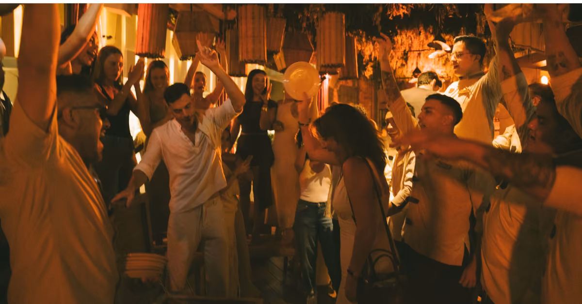 People dance and celebrate with raised hands and joyful faces at a festive Ilios Restaurant party under warm lighting.