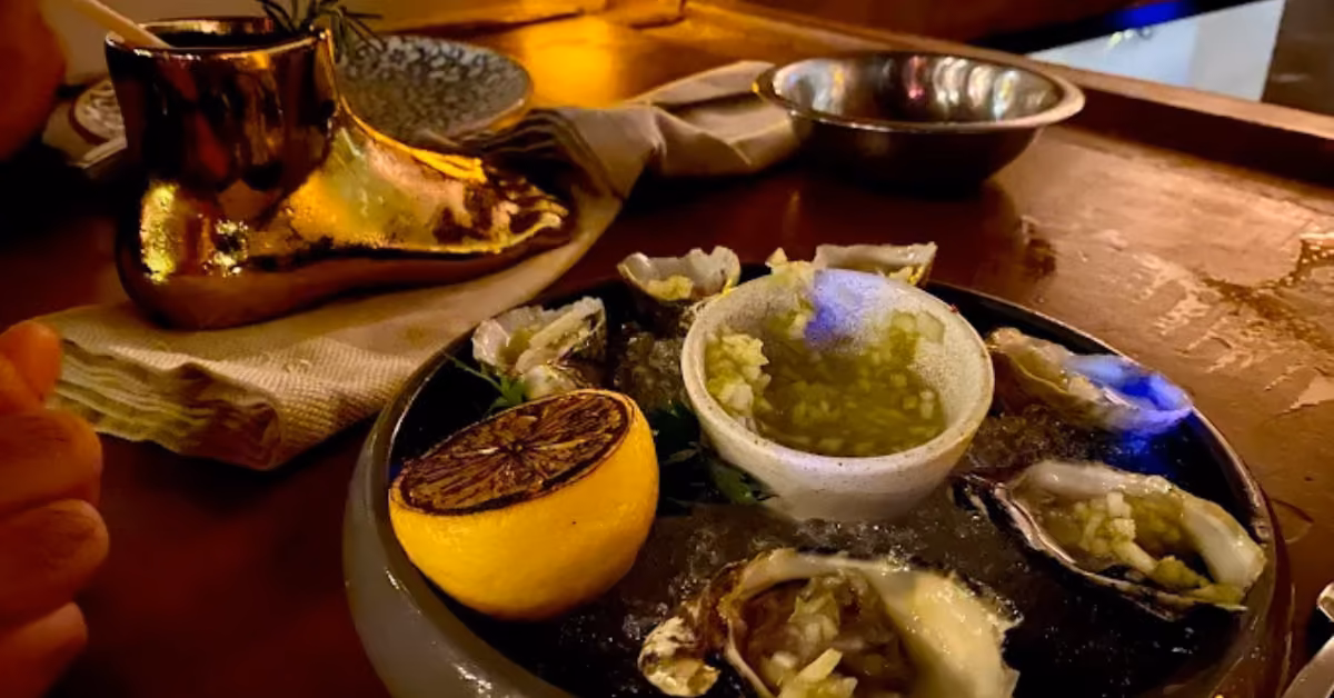 Fresh oysters on ice with grilled lemon and green sauce, served on a wooden table with a gold cup and cloth napkin.