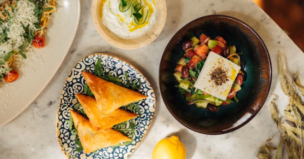 Mediterranean dishes on marble: spanakopita, Greek salad with feta, tzatziki, and a lemon arranged at Ilios Restaurant.