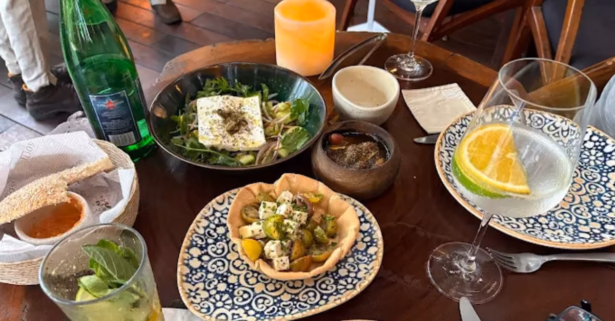 Dining table with salad, herbed cheese cubes, bread and honey, a green drink, sparkling water, orange water, and candle.