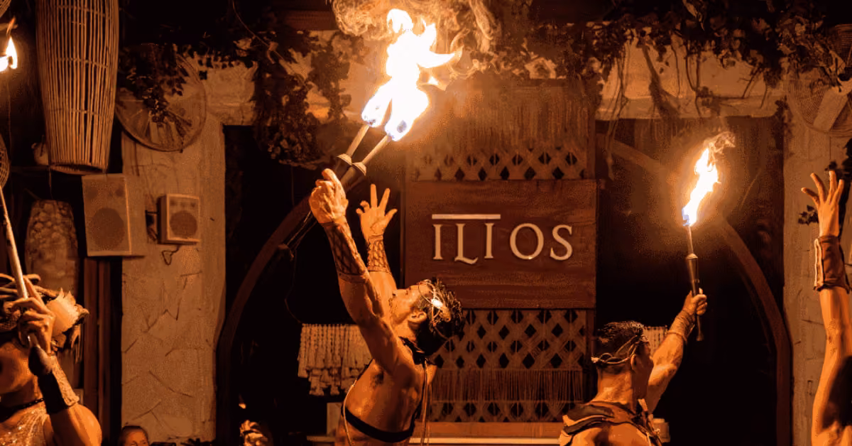Four performers juggle flaming torches at ILIOS Restaurant, arms raised under warm lights with a glowing ILIOS sign behind them.