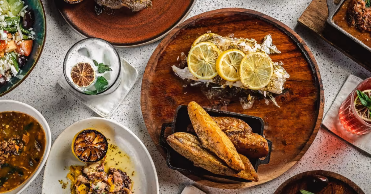Overhead view of a table at ilios restaurant with grilled fish, bread, salad, soup, cocktails, and assorted dishes.