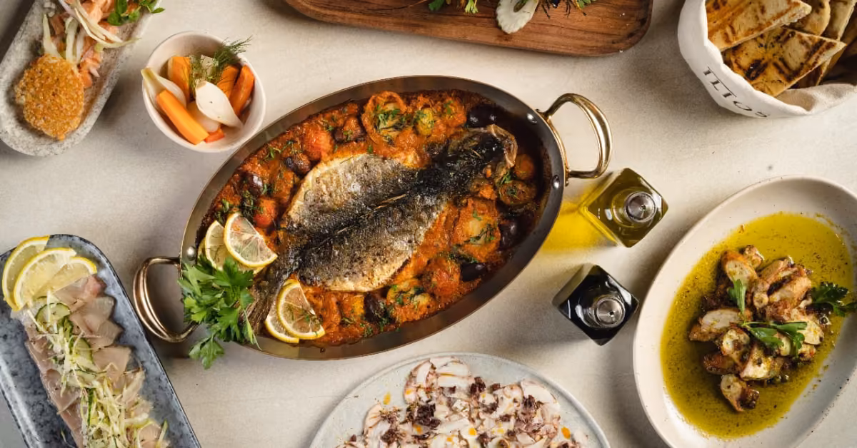 Mediterranean meal with baked fish in tomato sauce, artichokes, pickles, flatbread, olive oil, and small herb-garnished plates.
