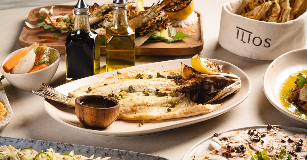 A table at Ilios Restaurant with Mediterranean fare: whole grilled fish, lemon, capers, bread basket, veggies, and appetizers.