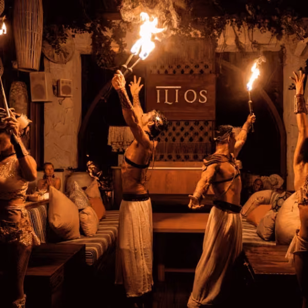Performers in white hold torches aloft in a lavish, dim lounge with cushions and an ILIOS sign as guests watch the fire show.
