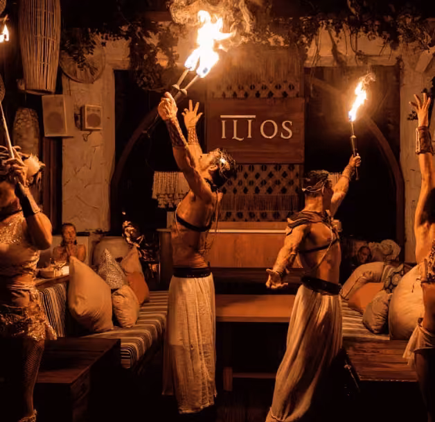 Performers in white hold torches aloft in a lavish, dim lounge with cushions and an ILIOS sign as guests watch the fire show.