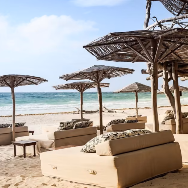 Beige lounge beds and wooden tables sit under thatched umbrellas on a sandy beach, facing a calm blue ocean and cloudy sky.