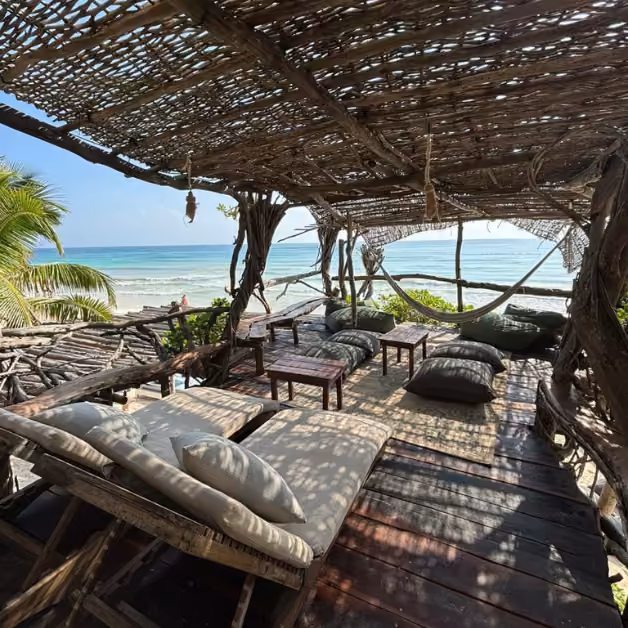 Shaded lounge with rustic wooden seats and cushions at Kanan Beach Club, overlooking a tropical beach and ocean under blue sky.