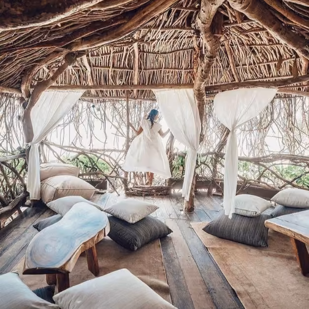 A woman in a white dress stands by an opening in a rustic treehouse with white curtains and cushions as sunlight streams in.