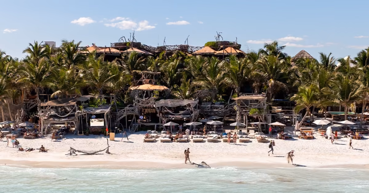 People relax by rustic wooden buildings and palm trees at a beachfront resort, strolling the white sand under sunny skies.