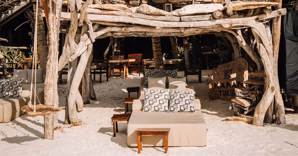 Wooden cabana with cushioned lounge chairs, patterned pillows, and a swing; tables and chairs in the background.
