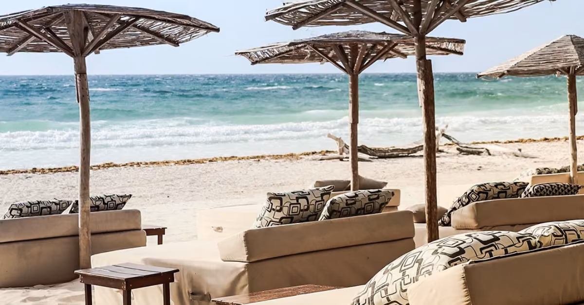 Patterned lounge chairs under straw umbrellas line the sandy shore, facing the ocean and gentle waves beneath a clear sky.