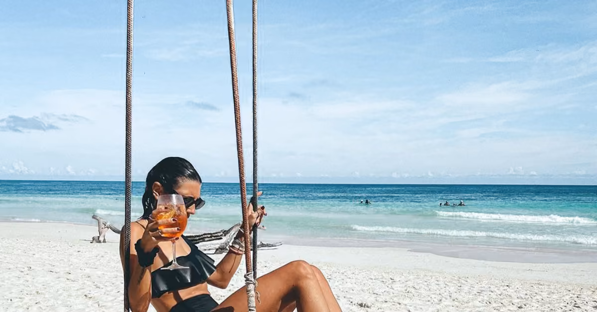 A woman in a black bikini and sunglasses sits on a swing at the beach, holding a drink with ocean and sky behind her.