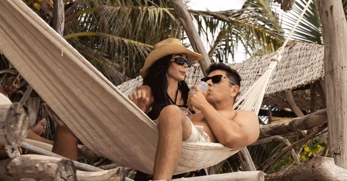 A woman in a sunhat and a man relax on a hammock at Kanan Beach Club, surrounded by palm trees; he sips from a bottle.