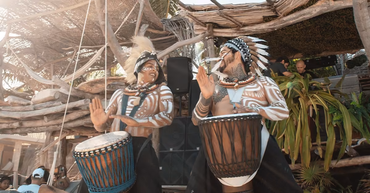 Two performers in tribal attire and body paint play large drums outdoors at Kanan Beach Club, moving energetically.