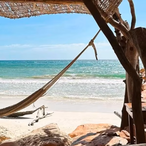 A hammock beneath a thatched palm shade at Kanan Beach Club overlooks sandy beach and turquoise sea under a bright blue sky.