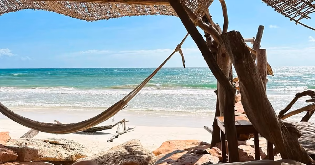 A hammock beneath a thatched palm shade at Kanan Beach Club overlooks sandy beach and turquoise sea under a bright blue sky.