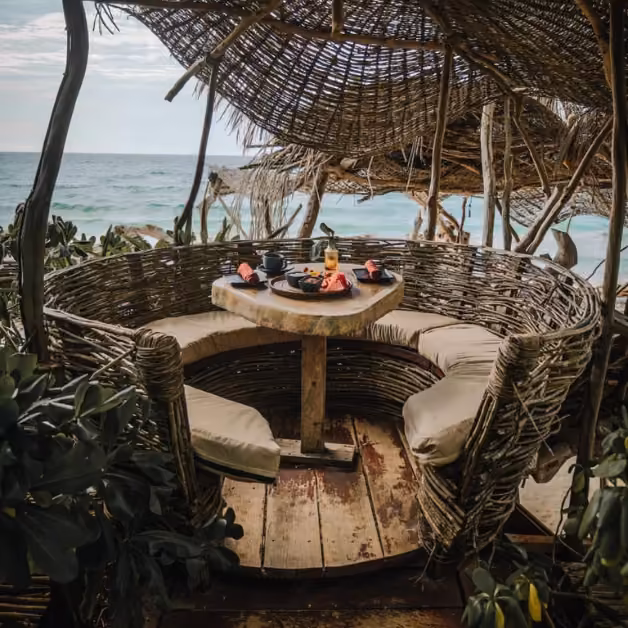 Round wicker seating with cushions and a wooden table set for dining at Kanan Beach Club, ocean view, lush greenery.