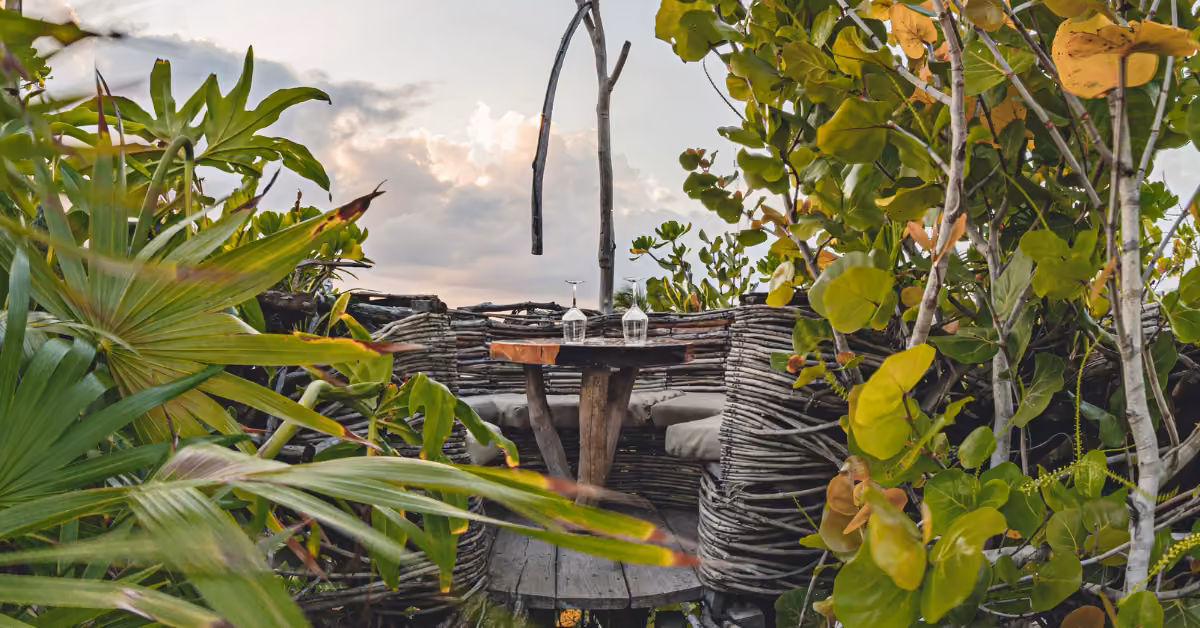 A rustic outdoor table with two wine glasses sits among lush greenery and wooden branches at Kanan Rooftop under cloudy skies.