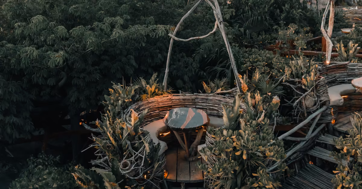 A round wooden table with two chairs sits on a raised terrace at Kanan Rooftop, surrounded by lush greenery and branches.