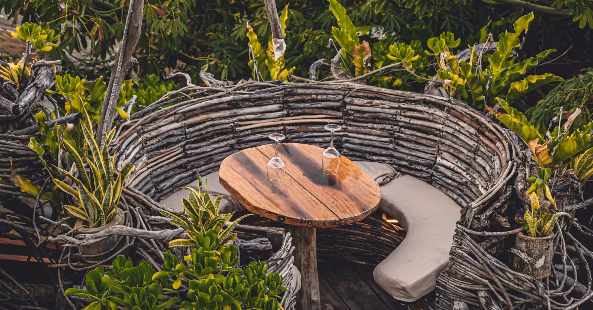 Cozy seating of woven branches encircles a wooden table with three upside-down wine glasses amid lush greenery on a rooftop.