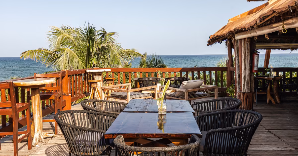 Wicker chairs and wood tables on Kanan Rooftop’s deck, ocean view, tropical plants, palm trees, and a clear sunny sky.