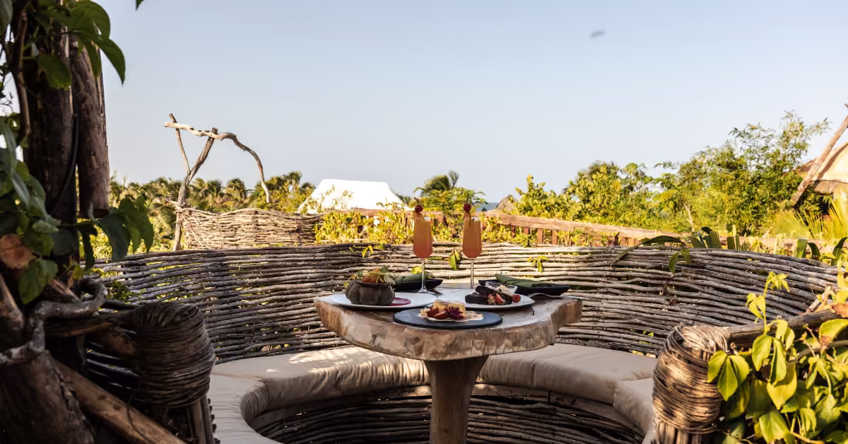 A cozy outdoor dining spot with a round wooden table, cushioned benches, food, drinks, and lush greenery on a sunny rooftop.