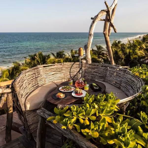 Cozy woven seating at Kanan Rooftop with a beach view, round table for two, dishes, drinks, and lush green plants around.