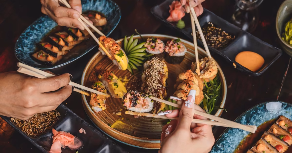 Three hands with chopsticks reach for sushi rolls on a round plate, surrounded by side dishes and sauces on a dark table.