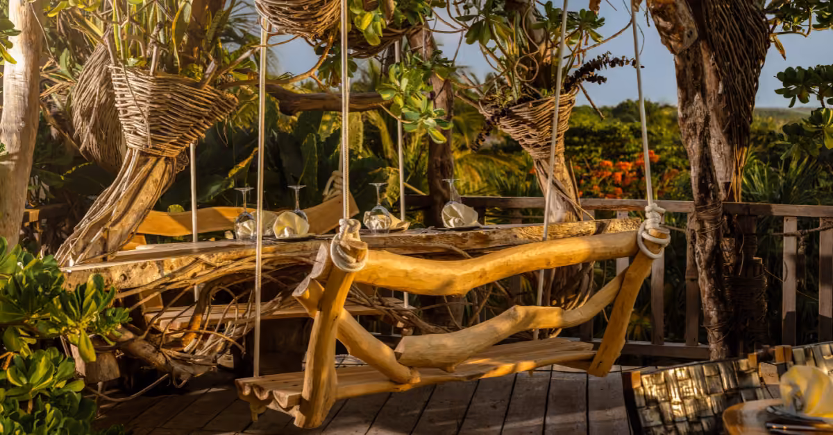 Karma Rooftop’s cozy dining area has a wooden swing seat, table set for guests, lush greenery, and wicker accents in sunlight.
