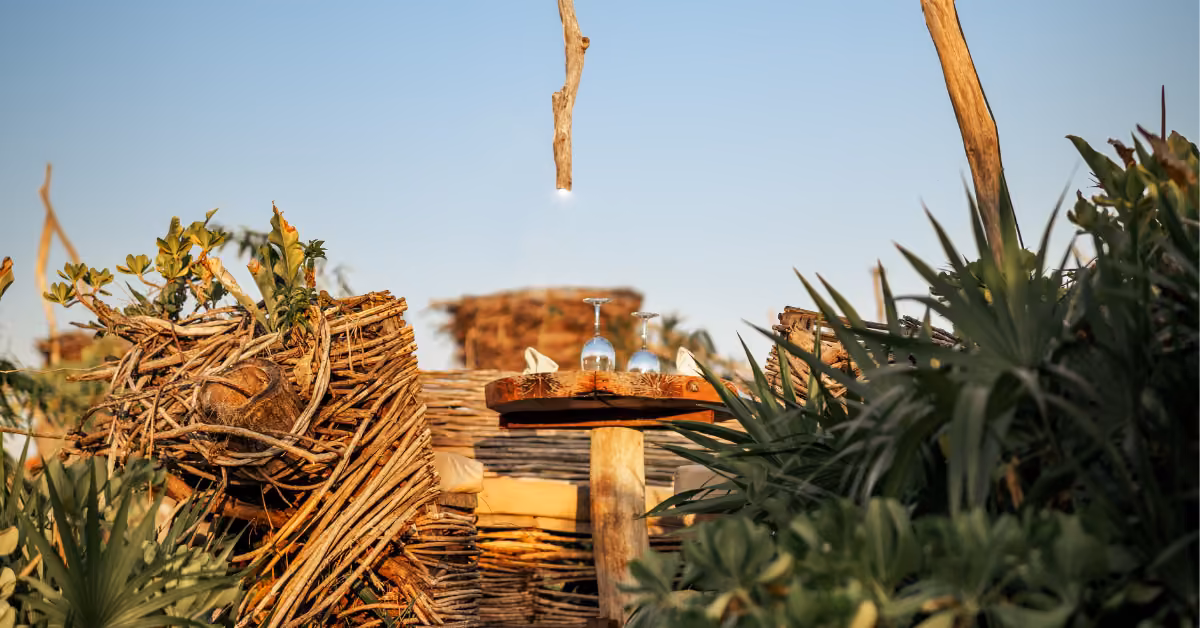 A wooden table with two upside-down glasses among woven branches and lush plants under a clear sky at Karma Rooftop.